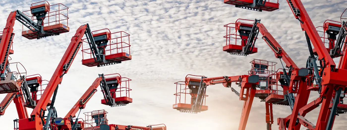 Multiple red aerial work platforms and boom lifts raised against a cloudy sky at an industrial equipment site.